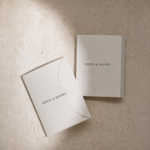 Minimal luxury hotel and resort stationery flat lay on a creamy textured stone surface with soft natural shadowing. A tilted curved-flap card in the foreground and a standing booklet in the background both feature elegant “HOTEL & RESORT” typography in black serif lettering.
