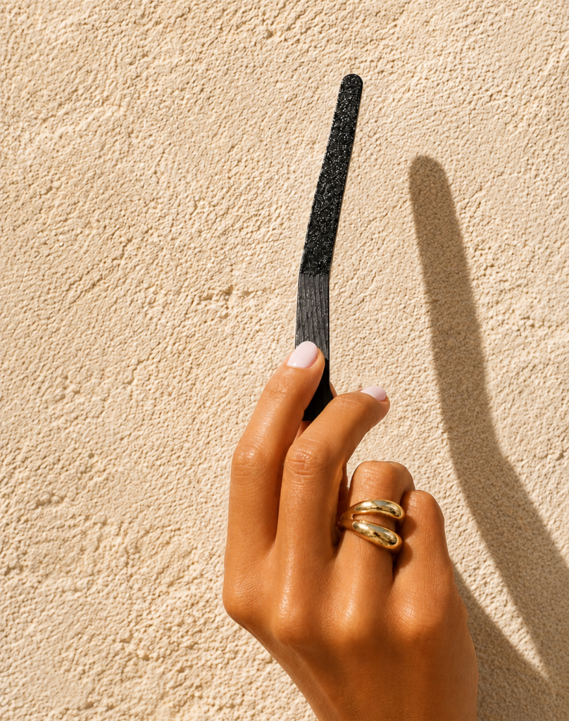 Close-up of a smooth tanned hand with gold rings holding a black textured scent strip against a creamy sandstone textured background with bright natural lighting and soft shadows.
