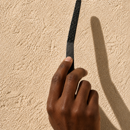 Close-up of a darker-skinned male hand holding a black textured scent strip against a warm sandstone wall with bright directional lighting and dramatic shadow detail.