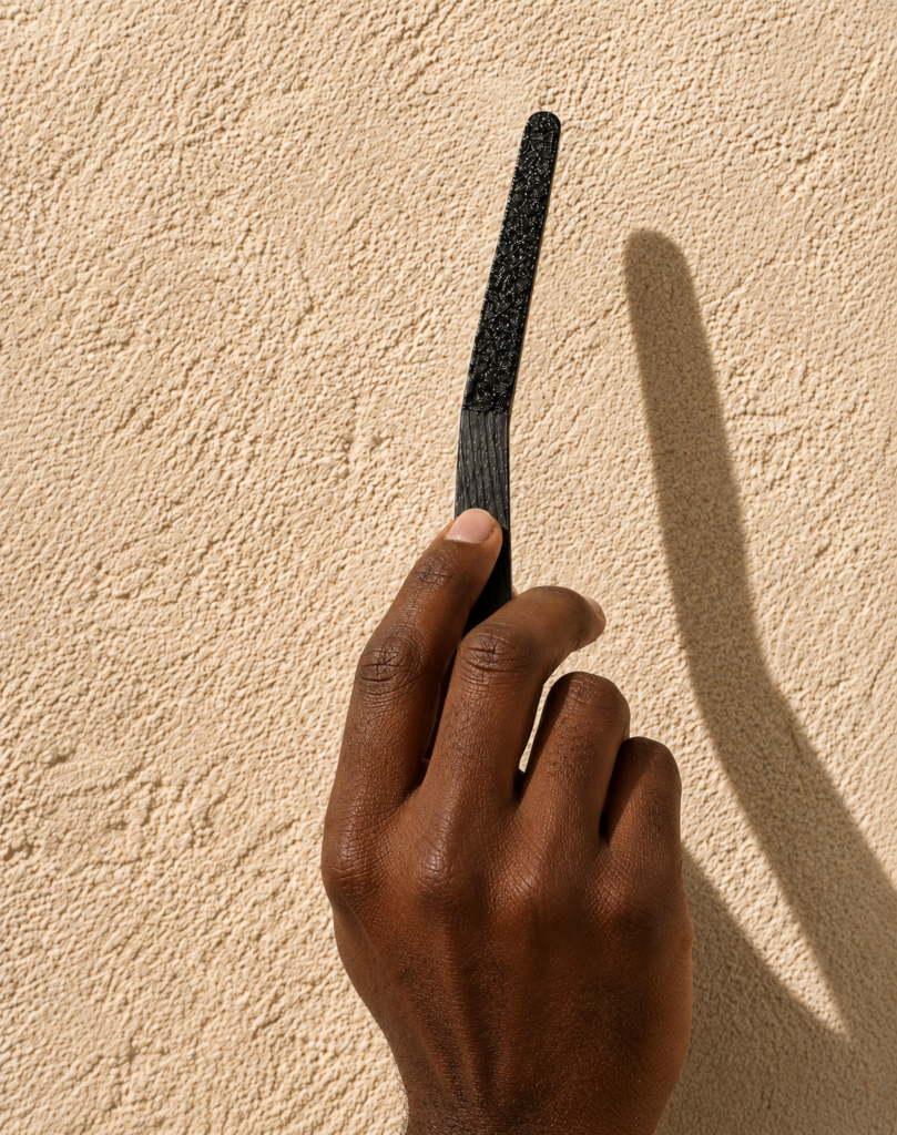 Close-up of a darker-skinned male hand holding a black textured scent strip against a warm sandstone wall with bright directional lighting and dramatic shadow detail.