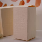 UX BOXES® | Leather-Touch Cream Straight Tuck End Gift Boxes™. Small cream-colored straight tuck end boxes embossed with a leather-like appearance against a watercolor swatch background in ambient lighting.