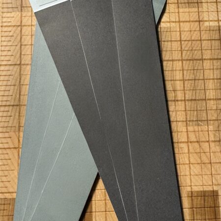 Mini envelopes by UX BOXES®. Six envelopes are laid flat over a wooden cutting board. Of the six, three dark gray envelopes are fanned overlapping a set of three green fanned envelopes. Self-seals are visible from the dark gray envelopes.