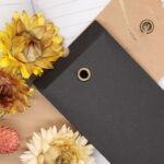 A black and kraft envelope atop a journal adorned with dried yellow flowers in bright lighting. The kraft envelope features a brass eyelet where brown twine is looped through.