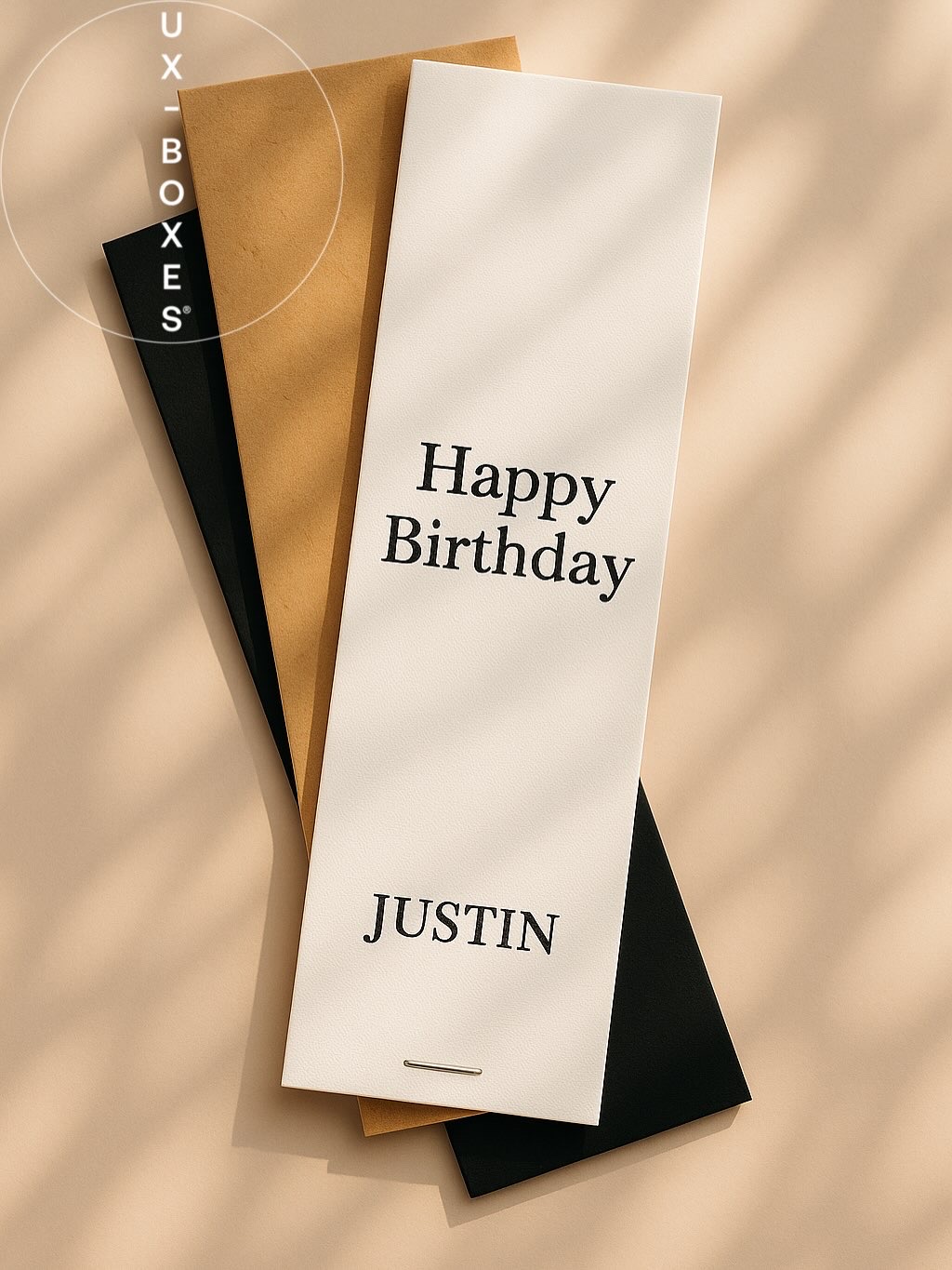 UX BOXES Trademark | Three sun-dappled overlapped envelopes laying flat on a light beige surface. A white envelope in the front of the overlapped stack reads: Happy Birthday JUSTIN. Underneath the white envelope is a Kraft envelope and underneath the Kraft is a black envelope.