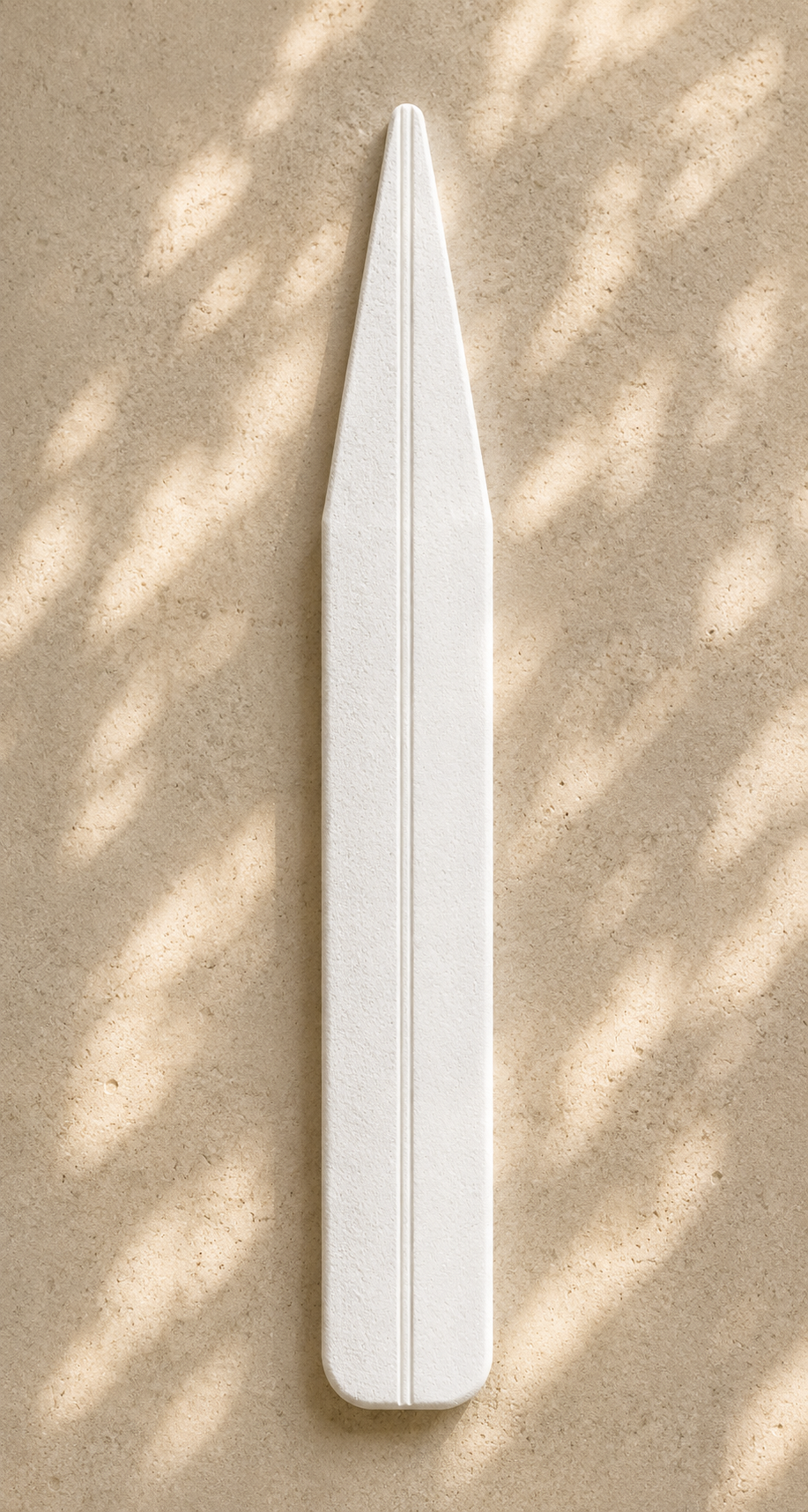 Minimal white perfume testing strip with angular tapered top, fine porous finish, and vertical groove on textured beige stone with organic sun dappling.