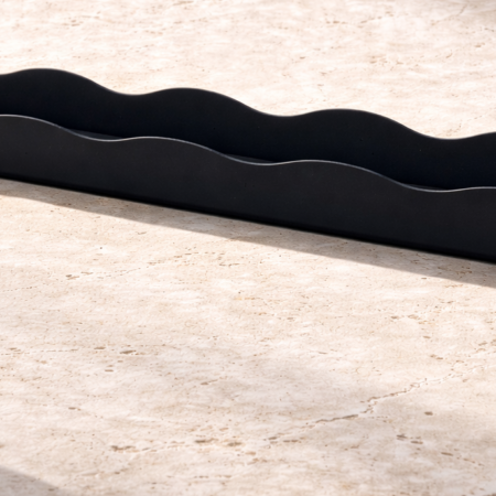 Close-up of a matte black wavy-edged tray resting on a cream-colored natural stone surface, with soft sunlight casting gentle shadows across the textured background.