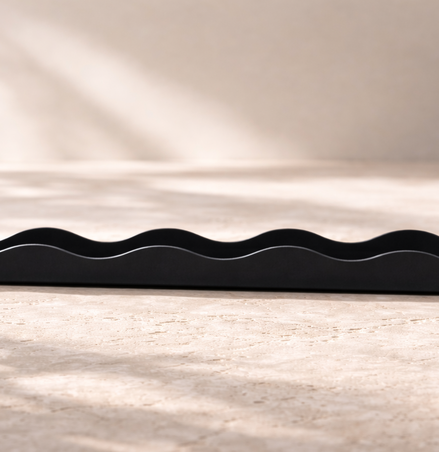 Straight-on side view of a slim matte black paper tray with smooth, undulating wave edges and rounded terminals, resting on a light beige stone surface with soft natural shadows in the background.