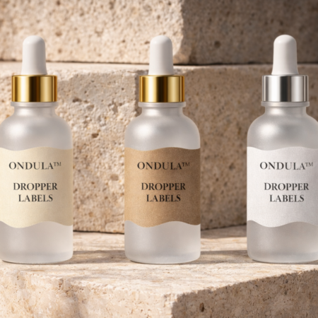 Three frosted glass dropper bottles with Ondula™ dropper labels in cream, kraft, and white linen textures displayed on a natural sandstone background.