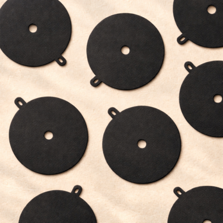 Flat lay of matte black round candle dust covers with centered wick holes and small arched pull-tab handles, arranged in a repeating pattern on a soft beige textured background.