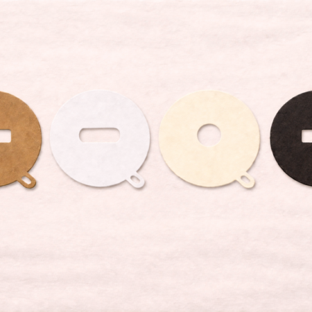 Four round textured cardstock candle dust covers arranged in a horizontal row on a soft off-white background. Each cover includes a small rounded pull tab at the bottom and features a different center cutout shape—rectangular, pill-shaped (rounded slot), circular, and rectangular—shown in kraft brown, bright white, soft cream, and matte black. Soft, even lighting highlights the subtle paper texture and clean die-cut edges.