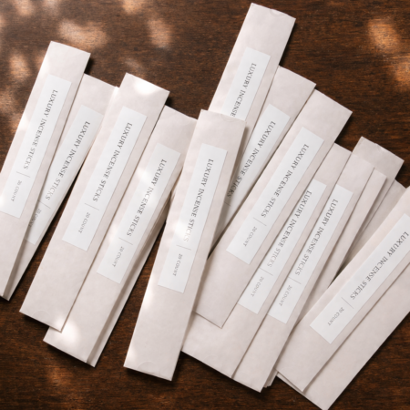 White incense sleeves arranged in a fanned layout on a dark wooden surface with soft sun dappling, each covered by a clean white label reading “LUXURY INCENSE STICKS | 20 COUNT” in an elegant serif style.