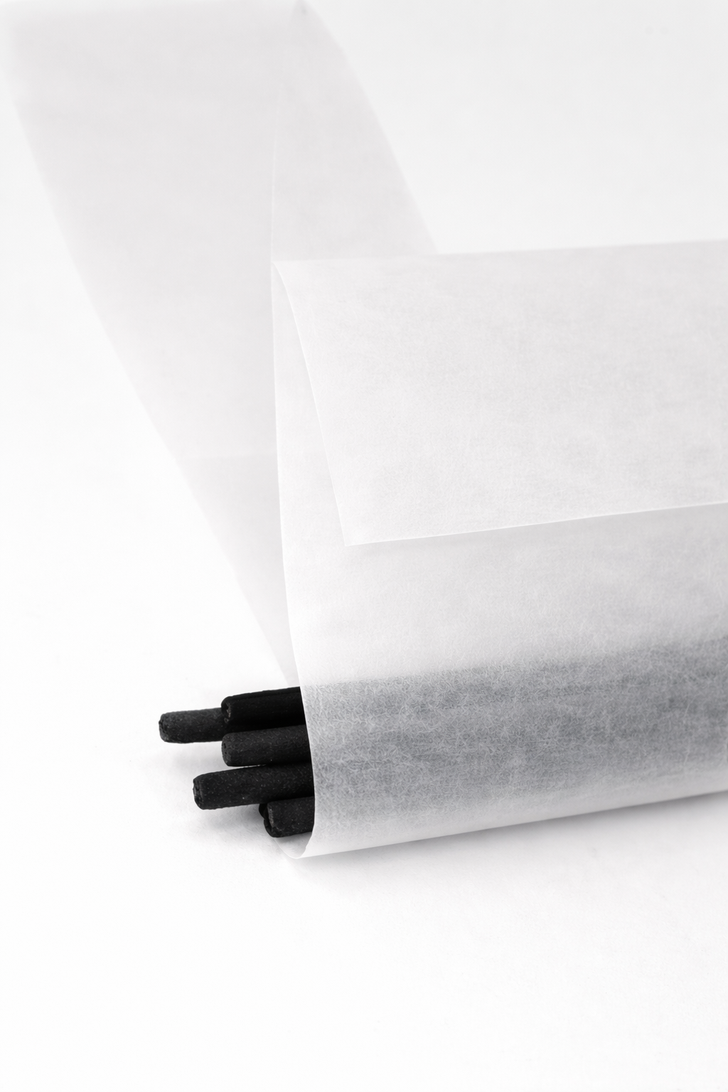 Bundle of black incense sticks partially wrapped in translucent vellum paper, with the frosted material softly revealing the sticks inside against a clean white background.