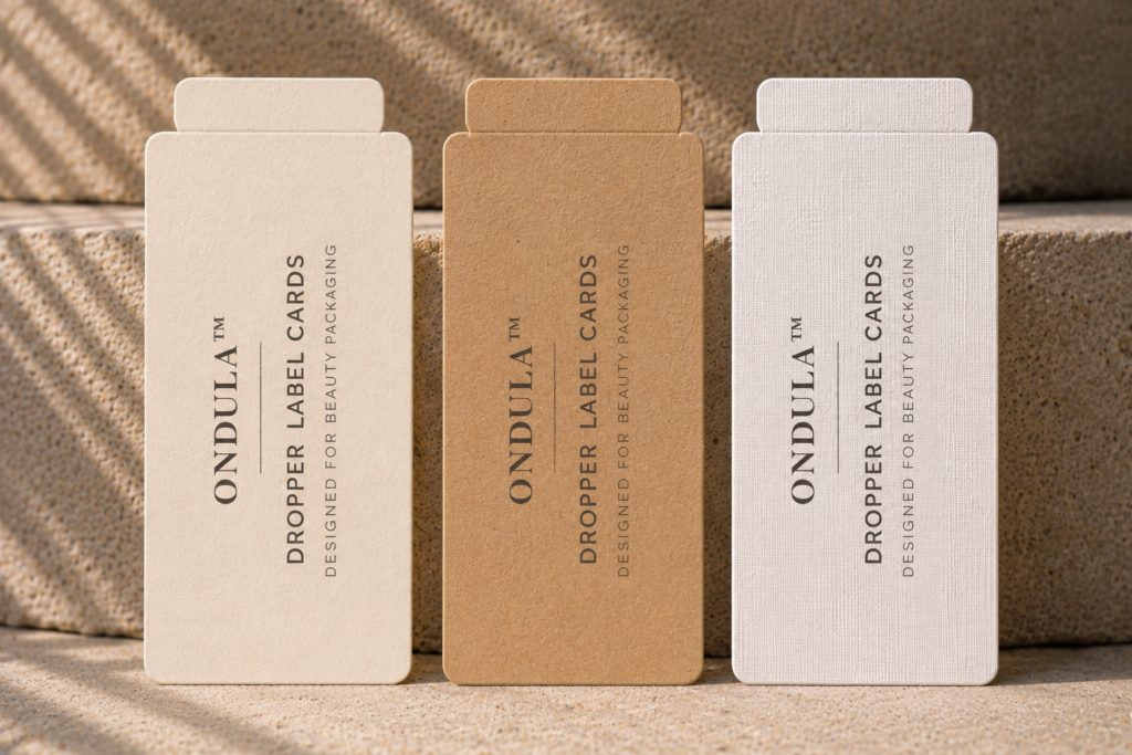 Three Ondula™ dropper label cards in cream, kraft, and white linen textured cardstock displayed against a natural stone background.
