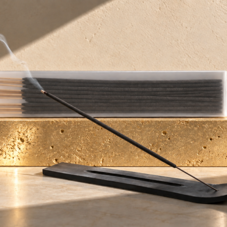 Black incense stick burning in a matte black holder beside packaged incense sticks displayed on a textured gold stone pedestal with stark directional lighting.