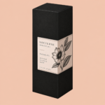 Matte black dropper box with sharp, dark gray fold lines and a small ivory label wrapping seamlessly across the front and side panels. The label features Nocturne Botanicals facial oil branding and delicate botanical illustrations. Presented in a three-quarter view against a soft peach background.