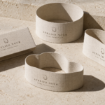 Luxury linen-textured belly bands in sculptural die-cut shapes with minimalist logo and typography, photographed on a creamstone background with stark directional lighting and dramatic shadows.